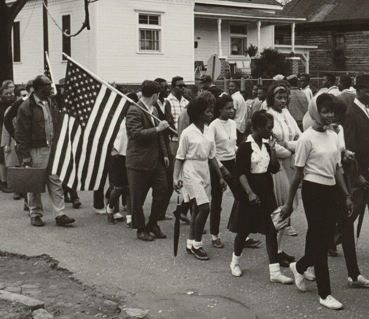 A group of civil rights marchers walk with an American Flag down a residential street in front of a white house.